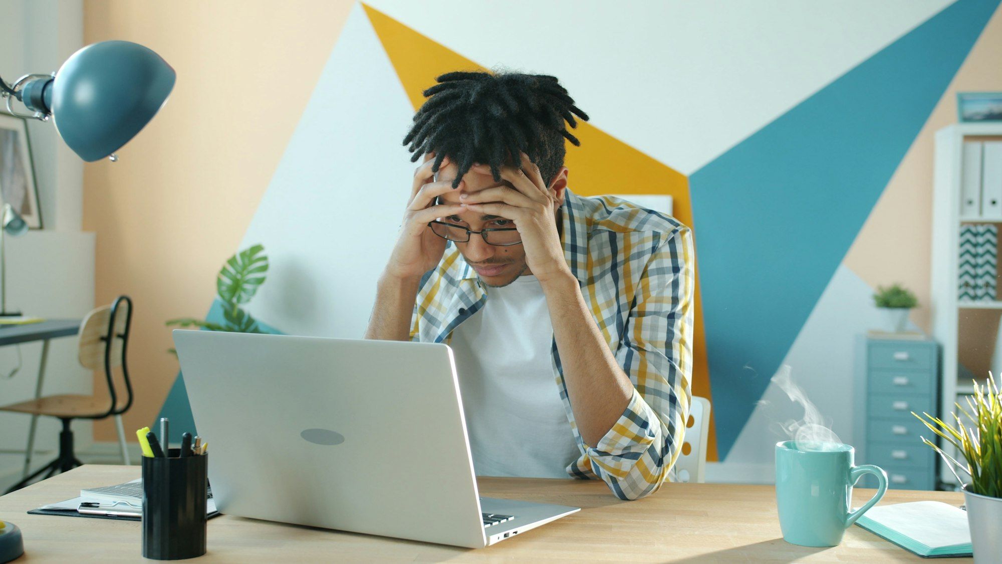 Business owner holding his head in frustration at his desk late in the day, laptop open and a hot drink beside him